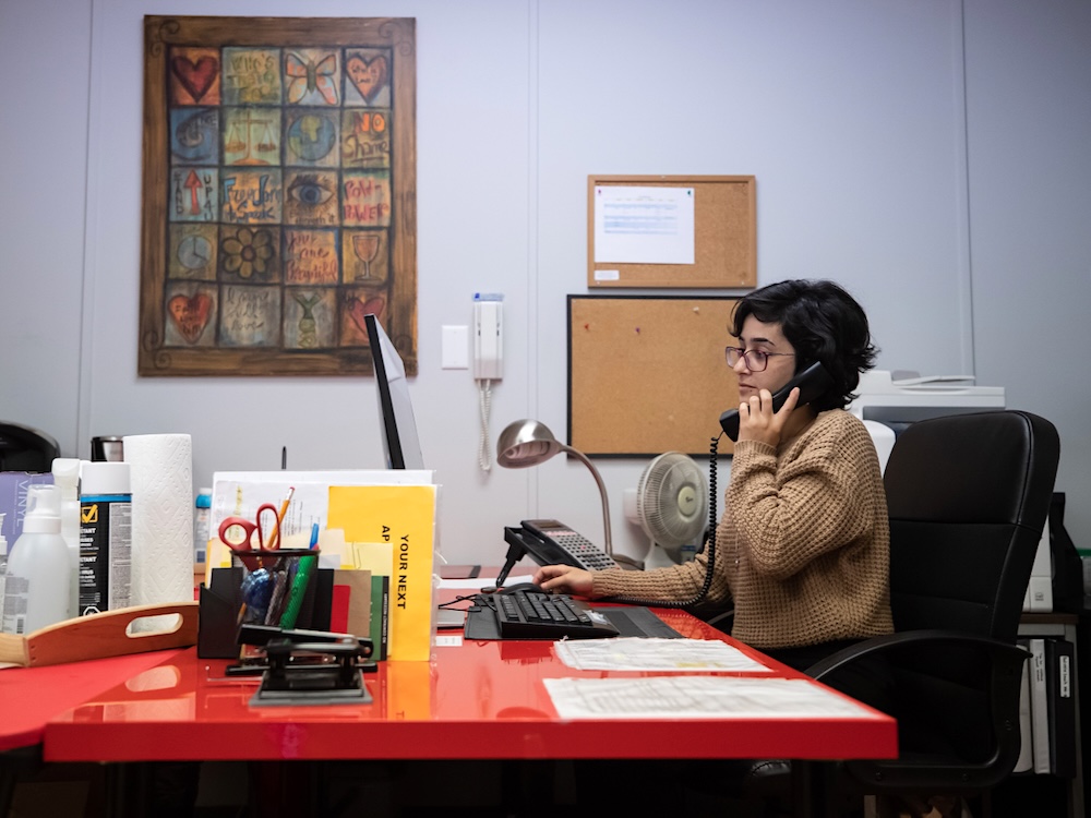 A woman sits at a desk in an office holding a landline phone receiver to her ear.