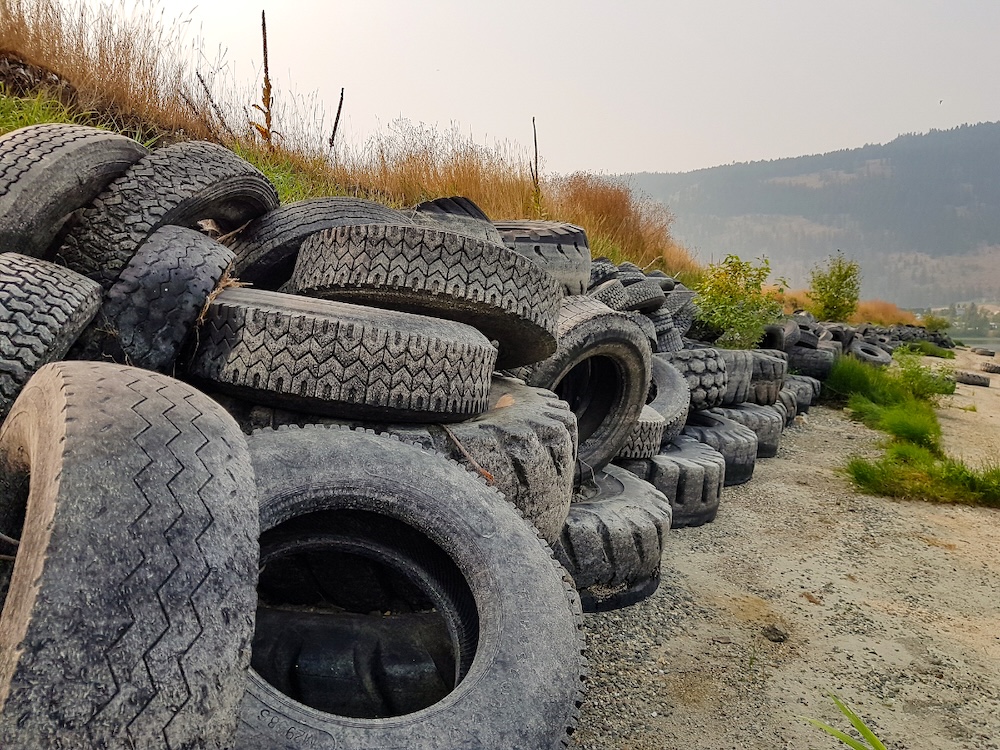 Large truck tires are piled several metres tall, separating a beach from a grassy field.