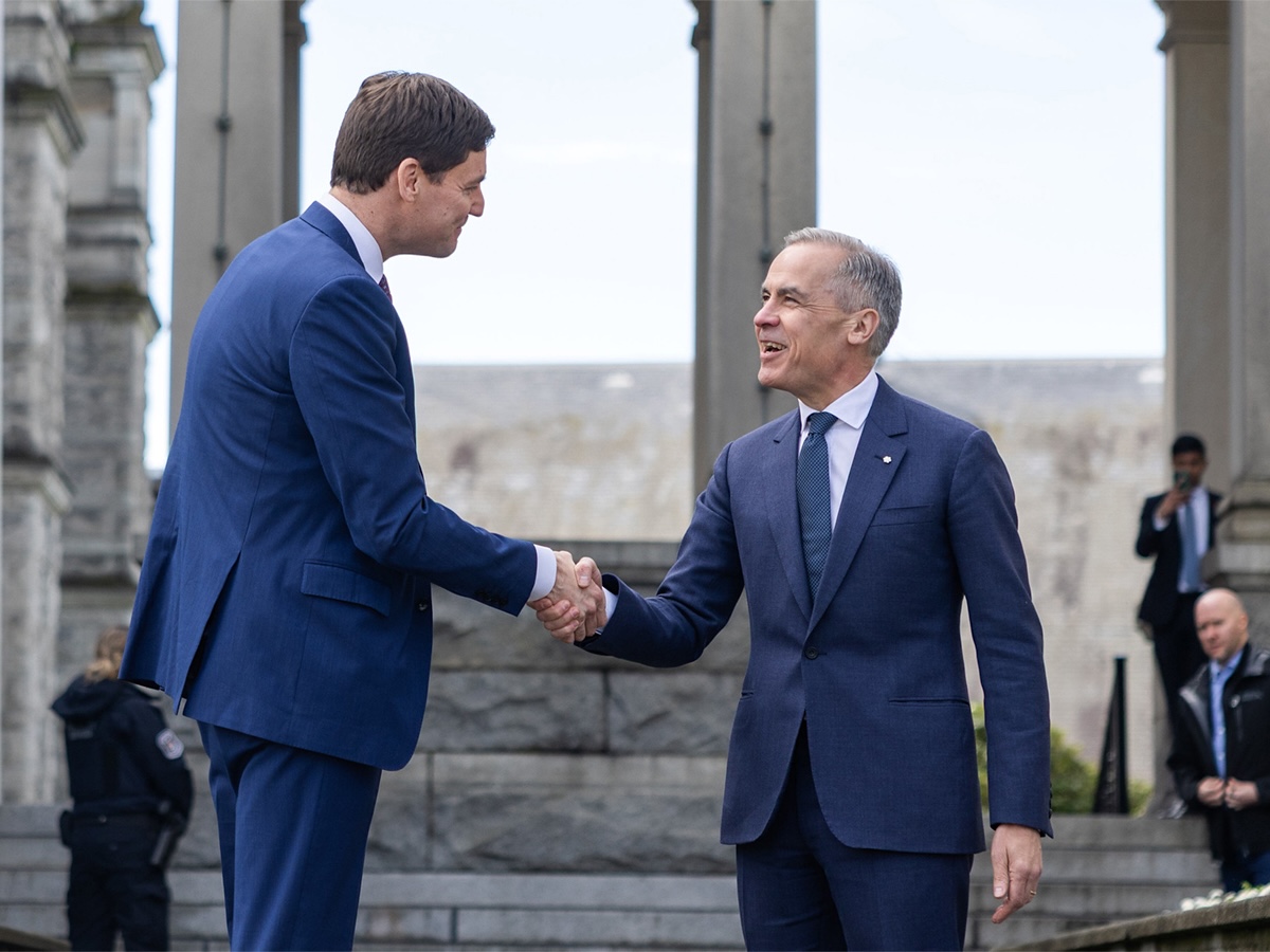 Two men in blue suits shake hands outdoors in front of a building with tall concrete columns.