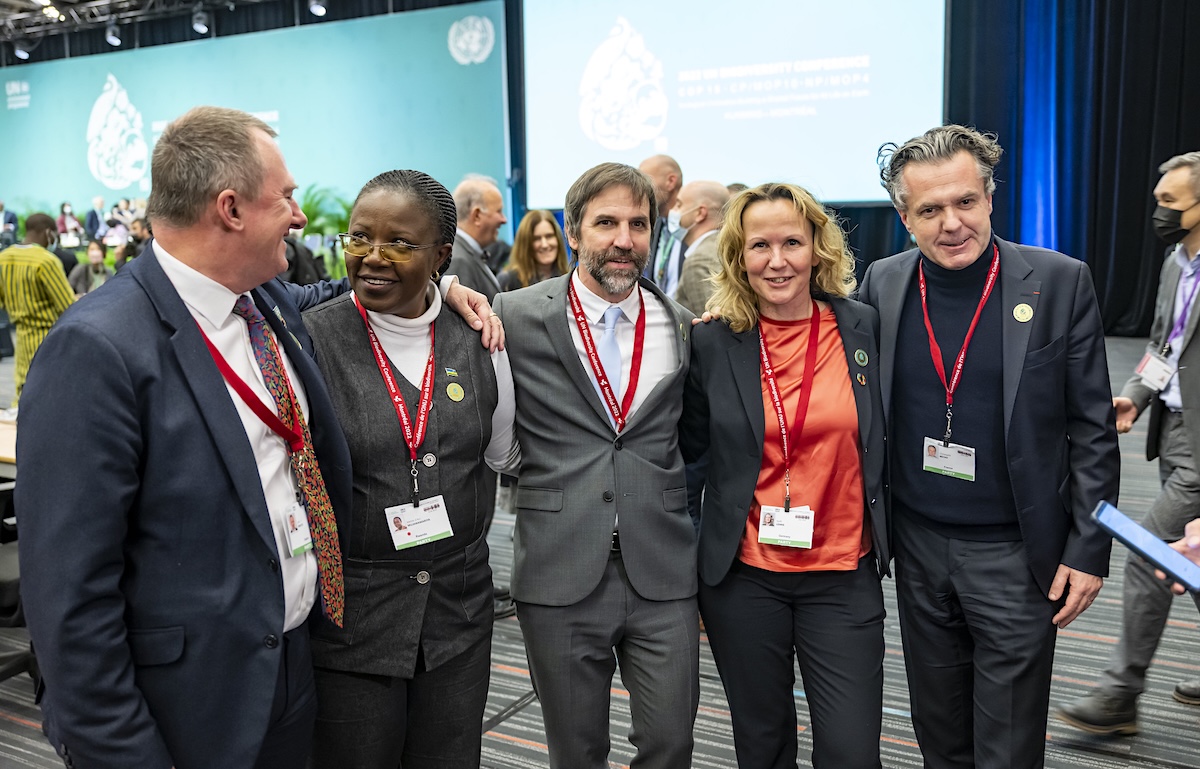 A group of five people wear suits and red lanyards, posing for a photo. Steven Guilbeault is at the centre.
