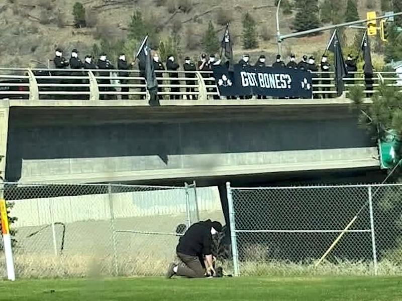 A group of men in black, wearing white masks, stands on a highway overpass holding flags and a white banner that says &lsquo;Got Bones?&rsquo; Another man kneels and pretends to dig in the grass below the overpass.