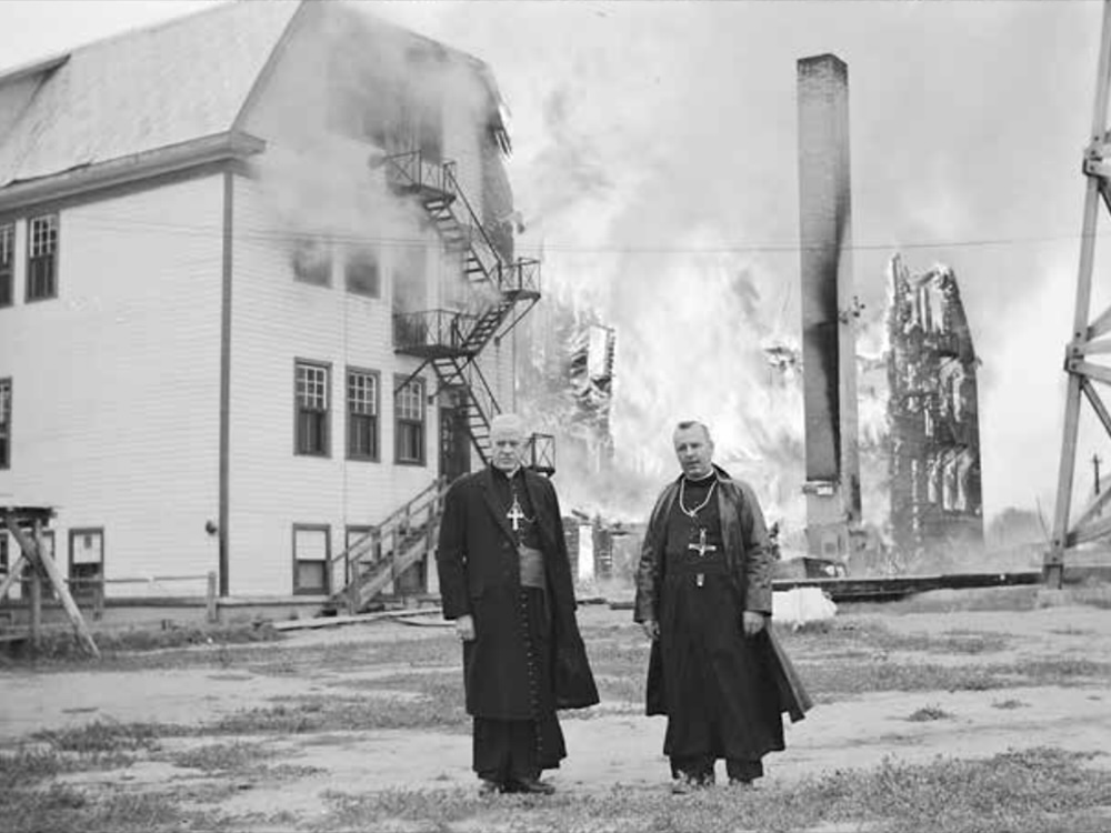 In a black and white historical photo, two priests stand in front of a burning three-storey building.