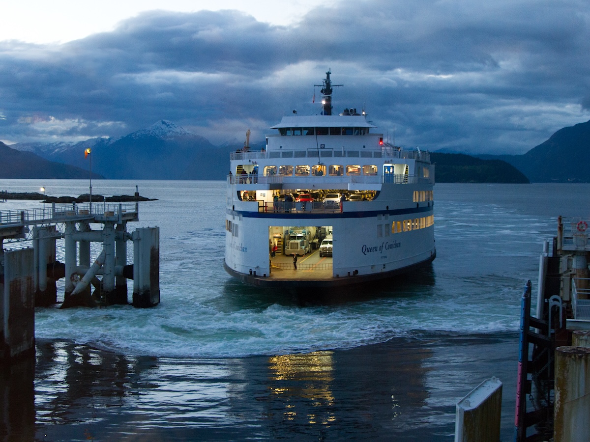 A BC Ferries vessel leaves a terminal under cloudy skies, with mountains in the background.