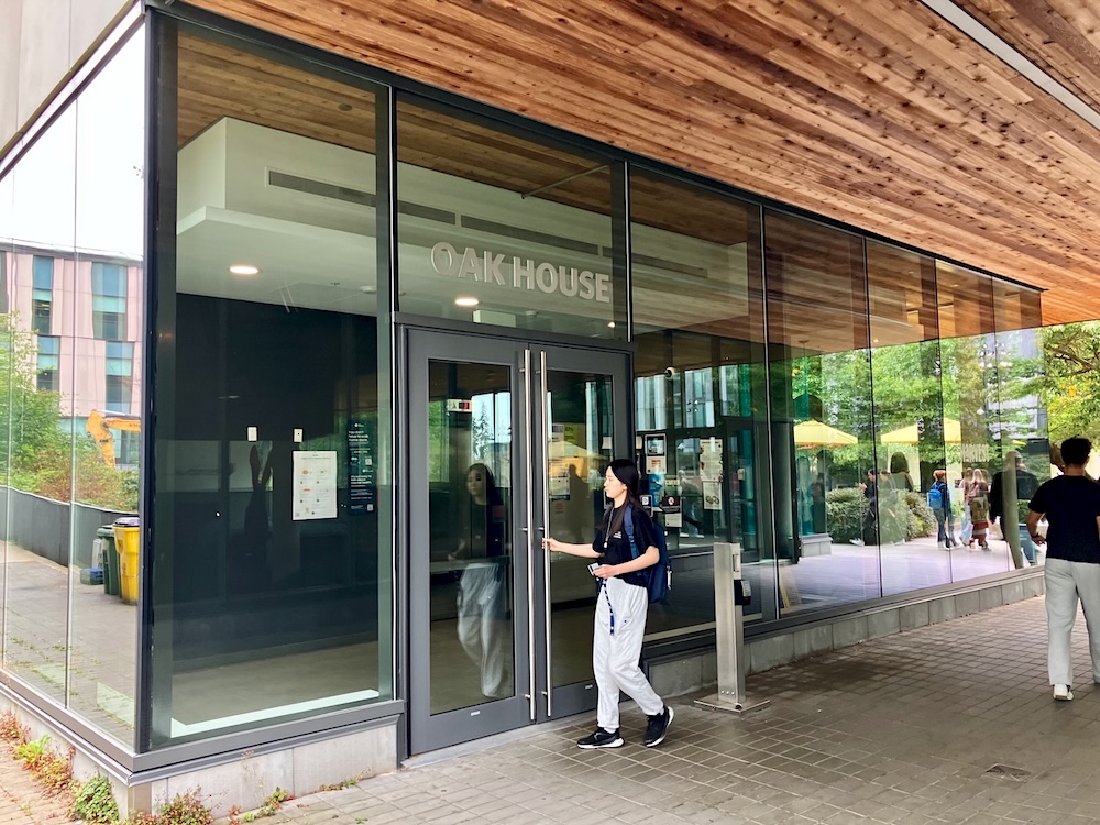 A student opens the glass door of a modern highrise. The sign &lsquo;Oak House&rsquo; can be seen above the door.