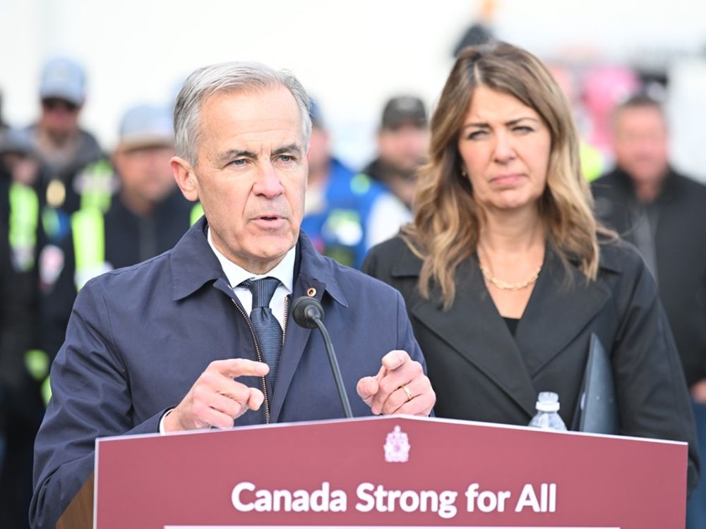 Danielle Smith, left, and Mark Carney sit at a table, smiling. There are Alberta flags and Canada flags behind them.