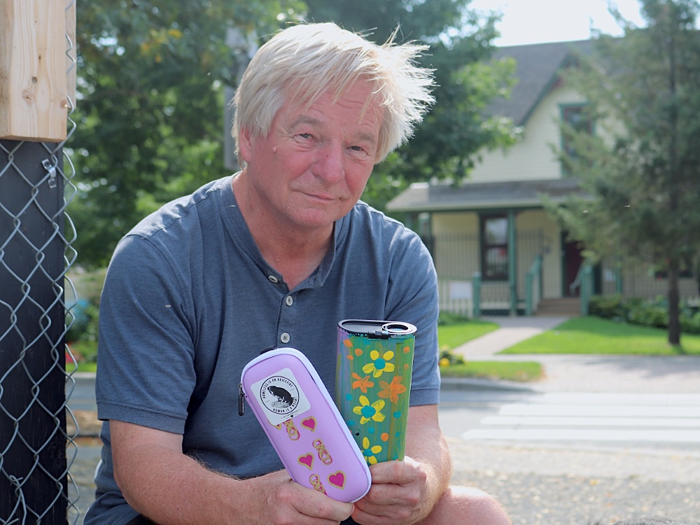 A man with blond-white hair holds two hand-decorated kits while sitting in front of a home and beside a chain-link fence.