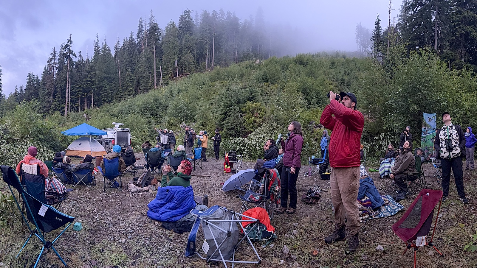 A large group of people stand and sit on lawn chairs, binoculars pointed at the sky.
