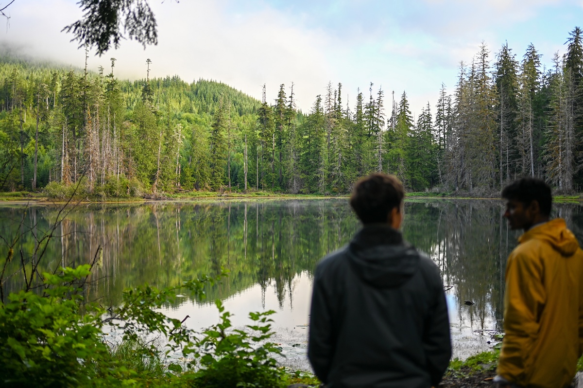 Two people stand on the side of a lake ringed with vibrant green trees.