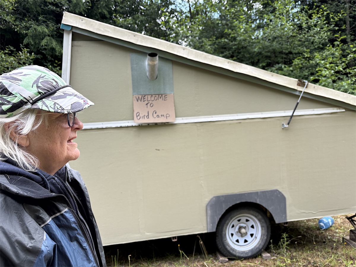 A woman wearing a baseball cap stands in front of her homemade trailer, which has a sign that reads, &lsquo;Welcome to Bird Camp.&rsquo;