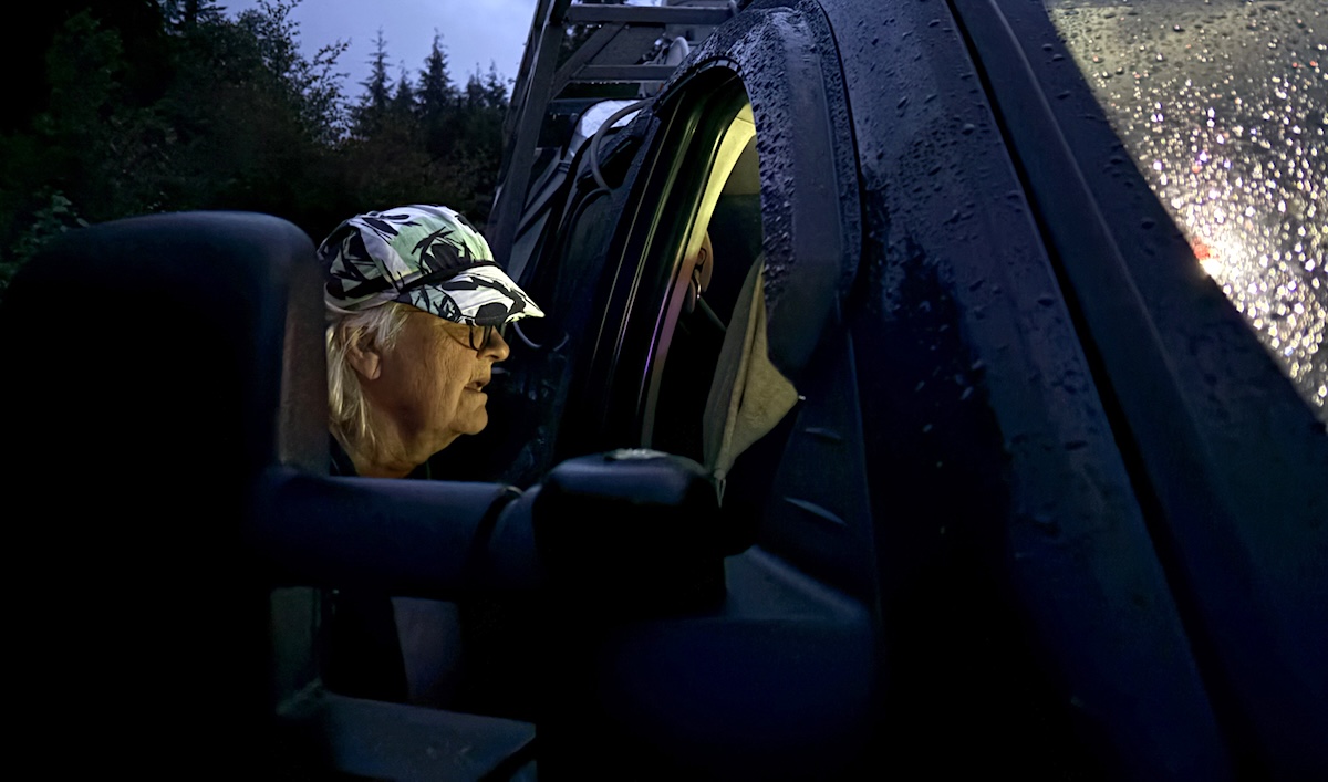 It&rsquo;s dark out. A woman wearing a baseball cap peers into a truck with its interior lights on.