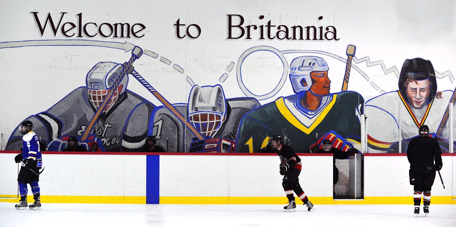 Three hockey players on the ice in a rink. Behind them is a large mural of hockey players with the words &lsquo;Welcome to Britannia.&rsquo;
