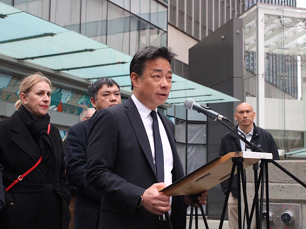 Ken Sim stands at a lectern and speaks into a microphone, with two people standing behind him.