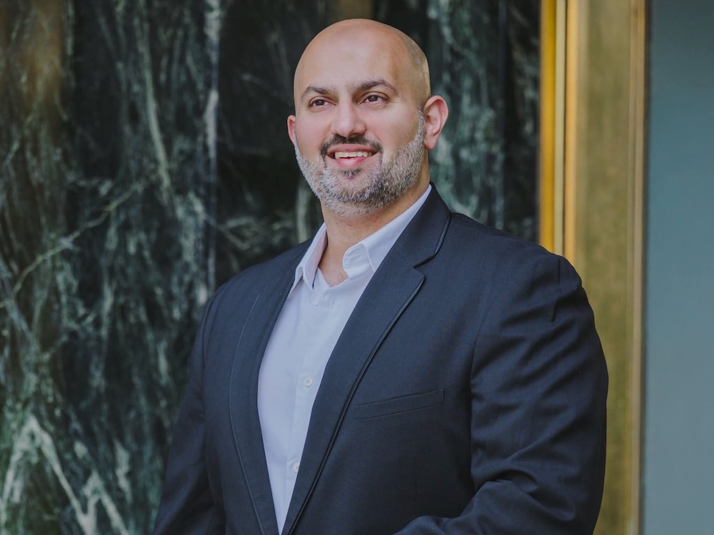 A man in his 30s who is bald with a grey beard and wearing a grey suit.