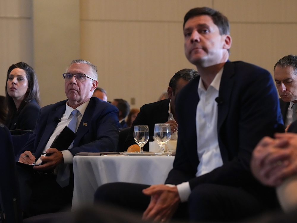 On the left, a man in his 60s with short grey hair wearing a suit leans back in a chair. On the right, a middle-aged man with short brown hair wearing a suit leans forward. The two men are sitting at a hotel conference room table.
