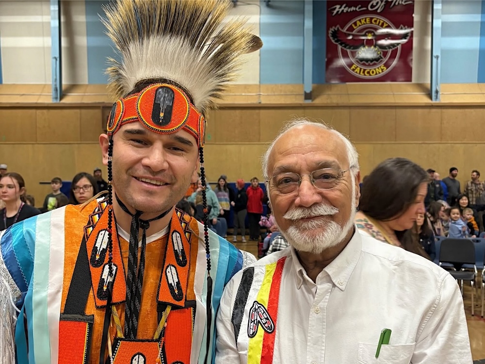 An Indigenous man wears a traditional headdress and orange Every Child Matters attire and stands next to an older man of South Asian descent wearing a white shirt with red, black, yellow and white stripes. The colours are from the Indigenous medicine wheel.