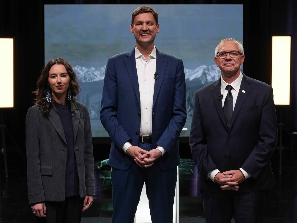 Three people stand beside each other. On the left is a 26-year-old woman in a dark jacket; in the centre a very tall man in a suit without a tie; and on the right an older man in a suit. 
