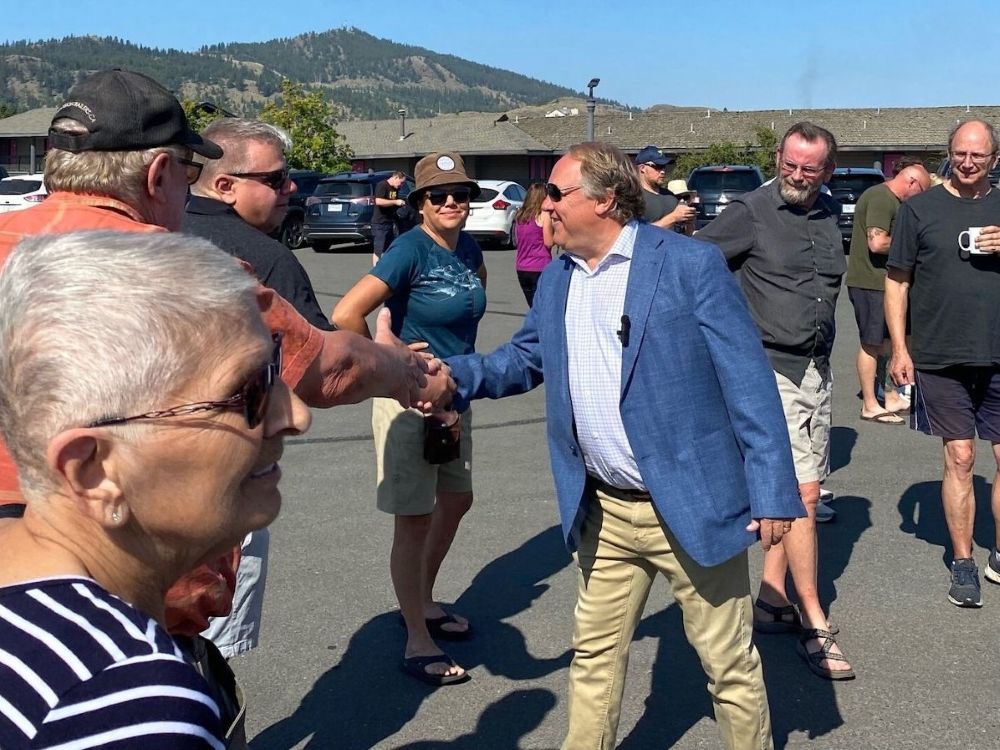A light skinned man in a blue jacket and khakis shakes hands with a group of people that surround him. In the background are hills covered in trees.