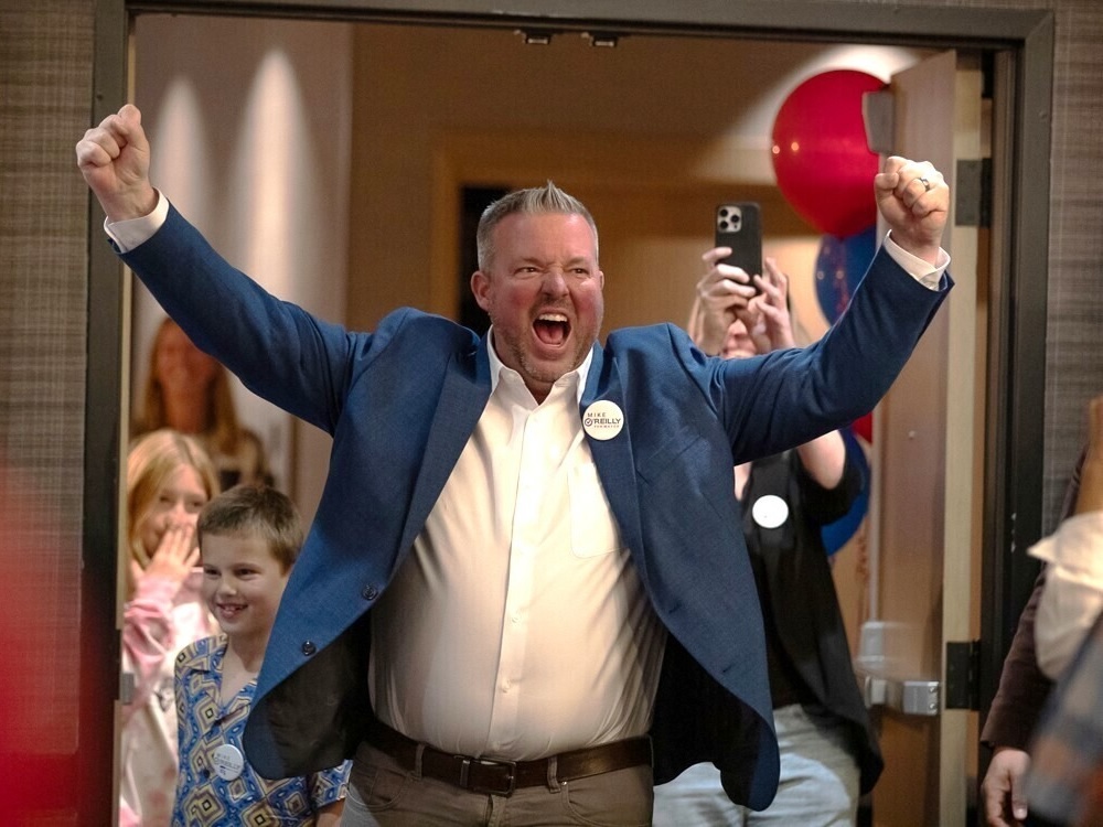 A light-skinned man in a blue blazer and white button-up shirt pumps his fists in celebration in a room full of people.