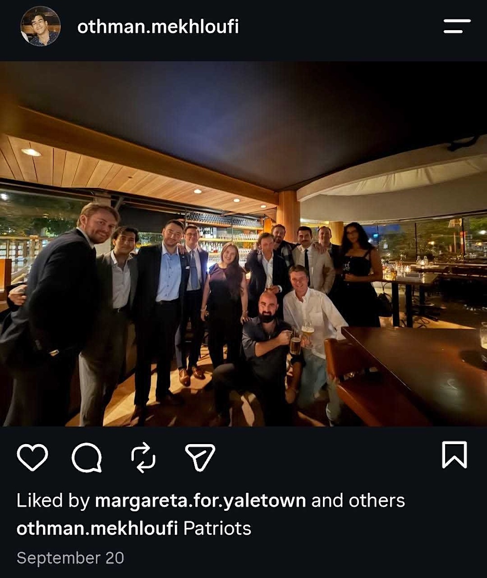A group of well-dressed people stand and kneel in a bar, some holding drinks.