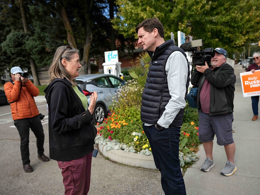 A tall man in his late 40s wearing a black puffy vest speaks to a middle-aged woman with shoulder-length blond hair on a small-town street.