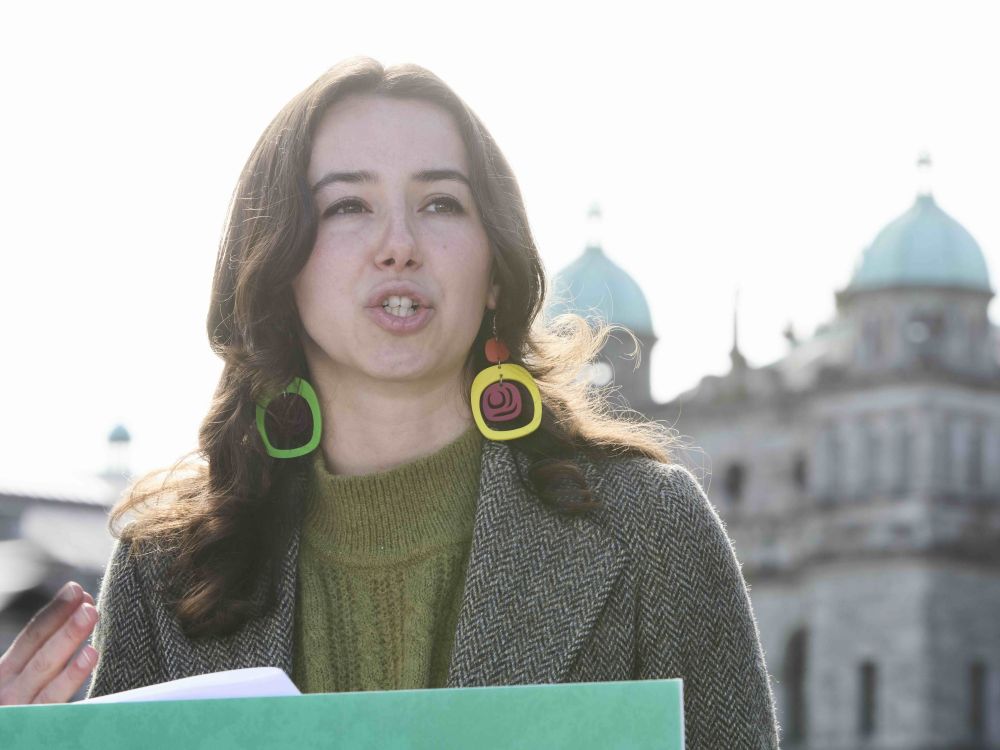 A young woman with long brown hair speaks at a podium with a historic gray stone building in the background.