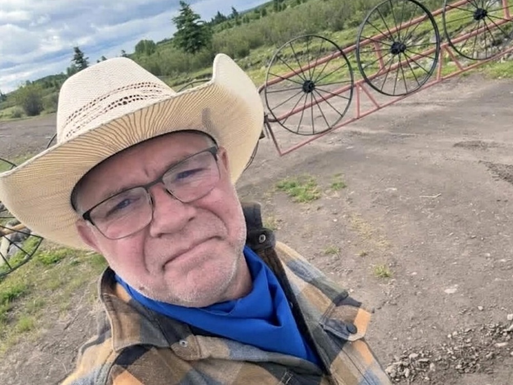 A middle-aged man wearing a straw cowboy hat takes a selfie in front of a gate made of old wagon wheels.
