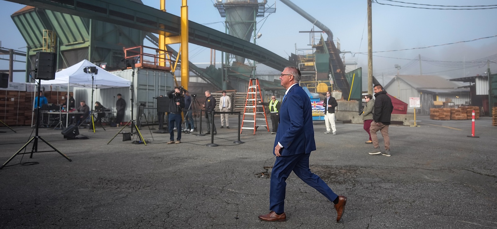 A middle-aged man in a blue suit walks through an industrial site.