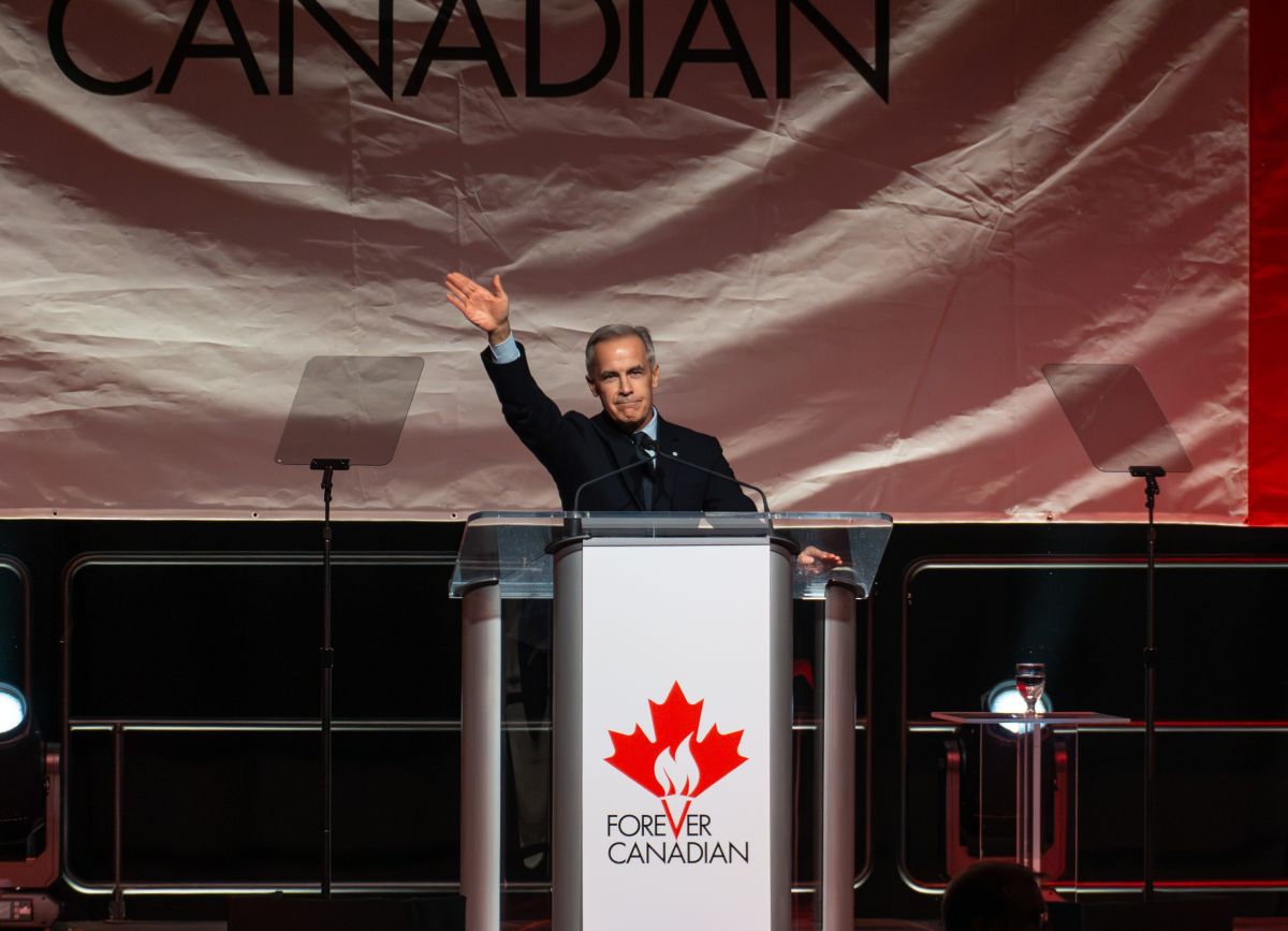 A middle-aged white man in a suit with a big maple leaf banner behind him stands at a podium and gestures.