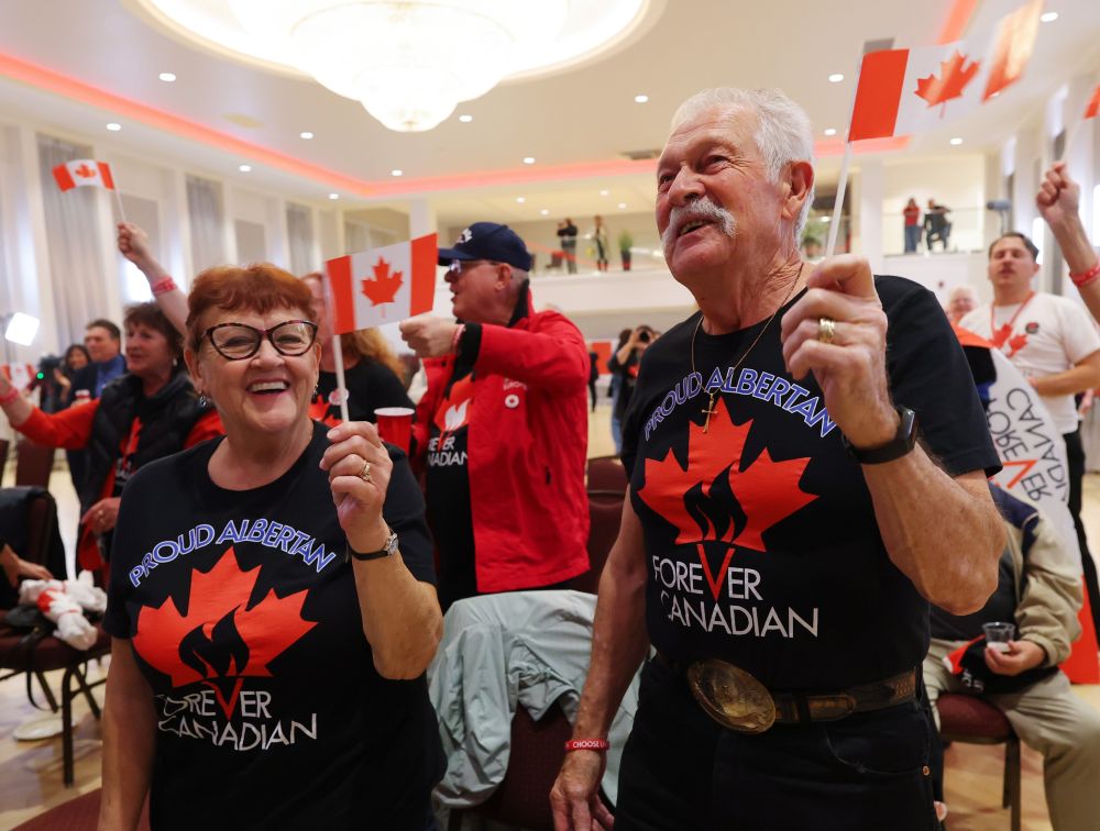 Two seniors, a woman and man, wearing shirts that say &lsquo;Proud Canadian,&rsquo; smile and wave Canadian flags in an ballroom with others gathered.