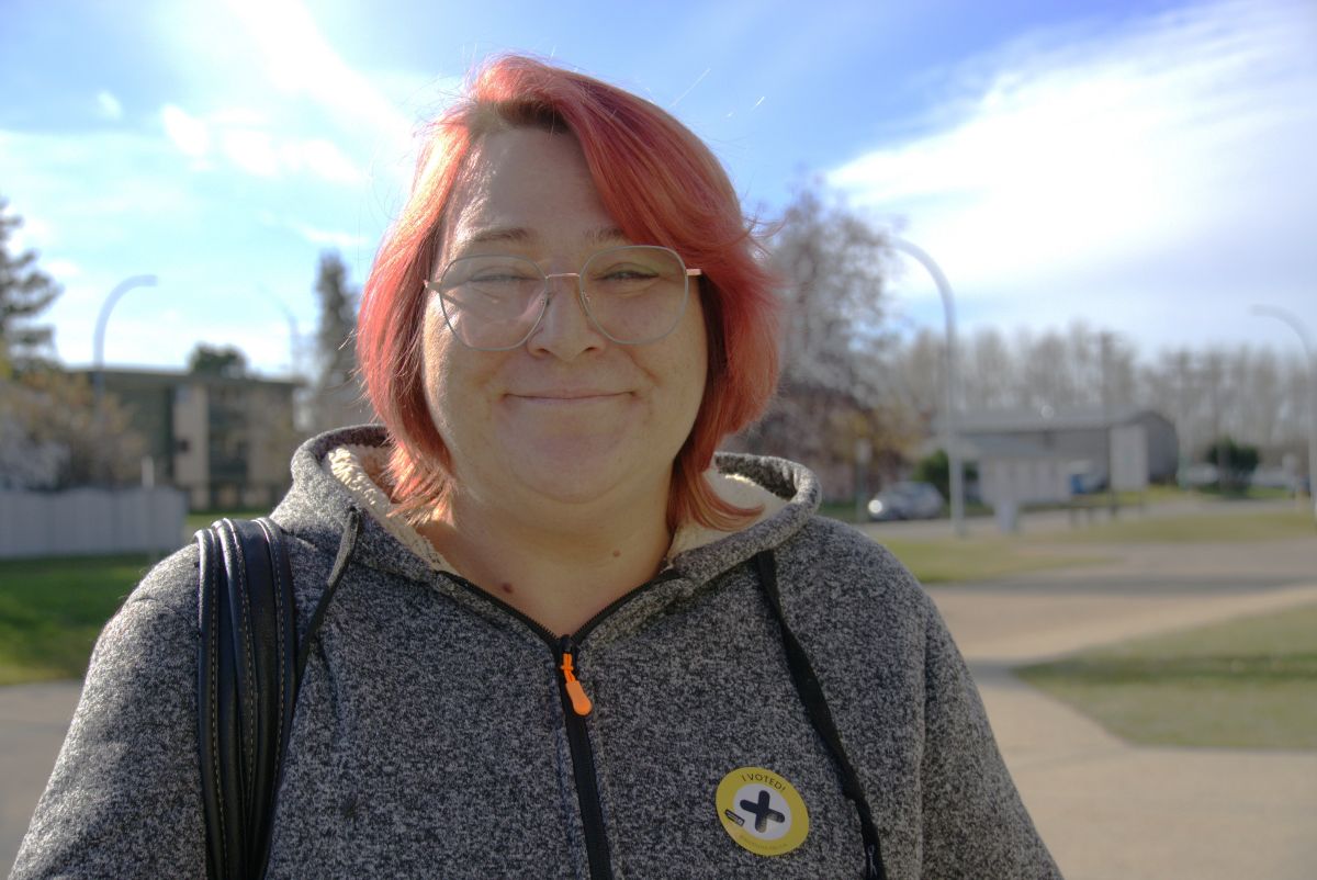 A light-skinned woman with red hair stands outdoors and smiles at the camera.