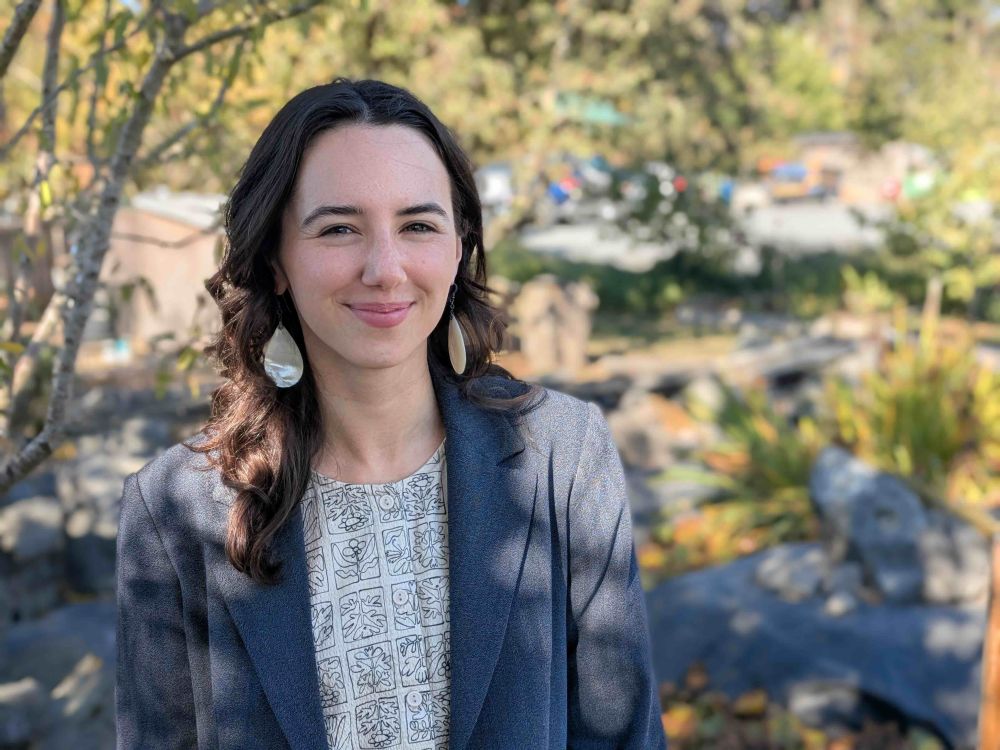 A 26-year-old light-skinned woman with long dark hair stands outdoors and looks at the camera. She wears large earrings and a suit jacket.