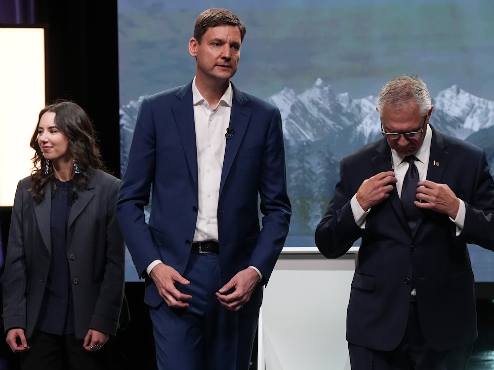 A young woman with long dark hair, a very tall middle-aged man and an older man stand awkwardly on a TV stage.