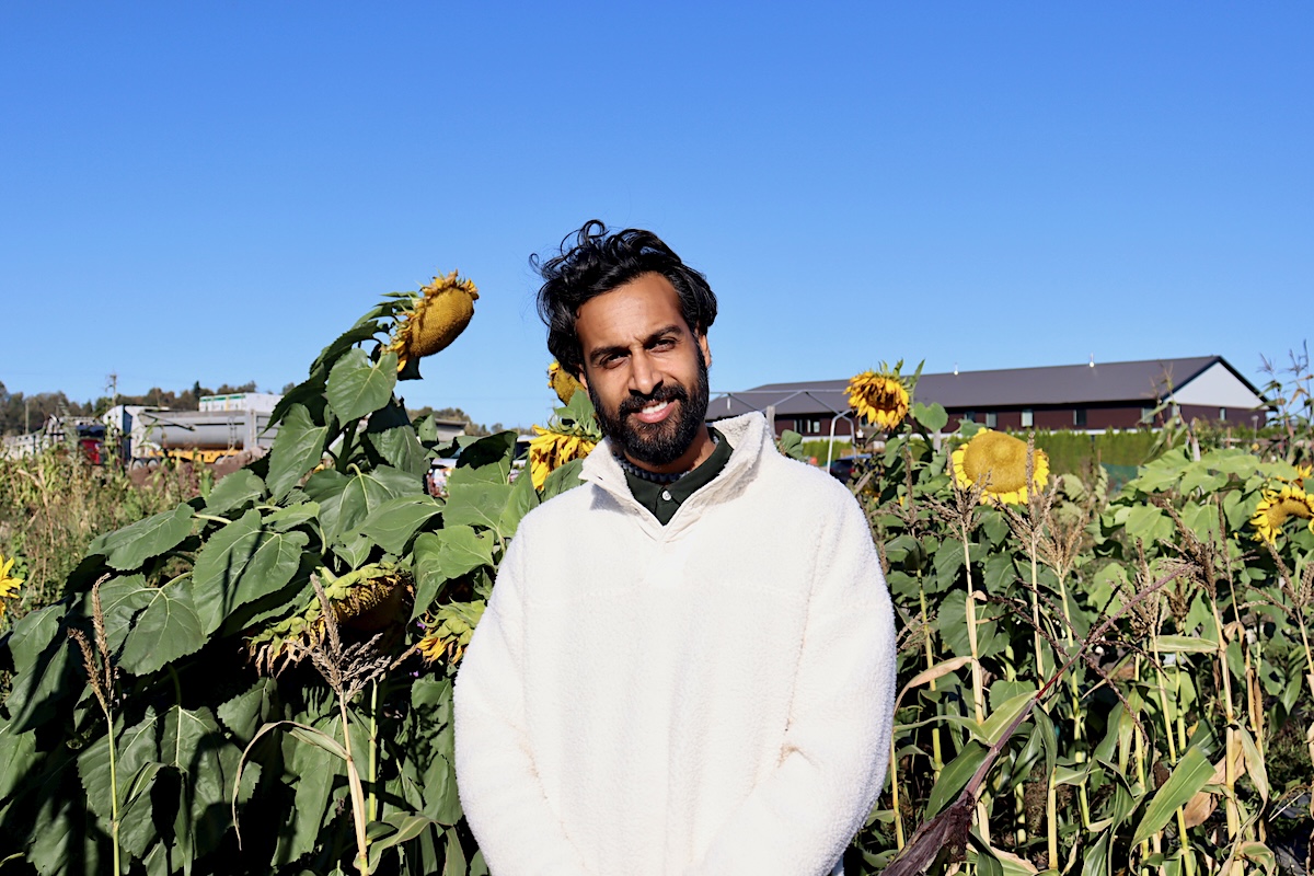 A man with shoulder-length black hair and a black beard, wearing a white sweater, stands in front of sunflowers on a farm.
