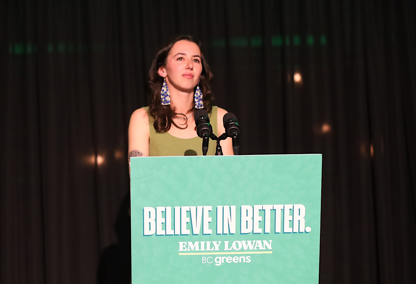 Emily Lowan, a 26-year-old light-skinned woman, stands at a lectern with a sign that says &lsquo;Believe in Better.&rsquo; 