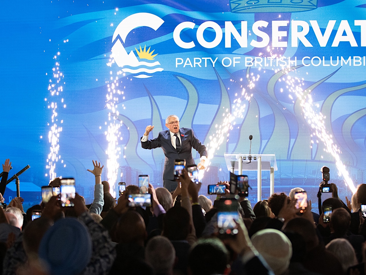 A white-haired man in a suit pumps his fist on a stage as pyrotechnics fire around him.