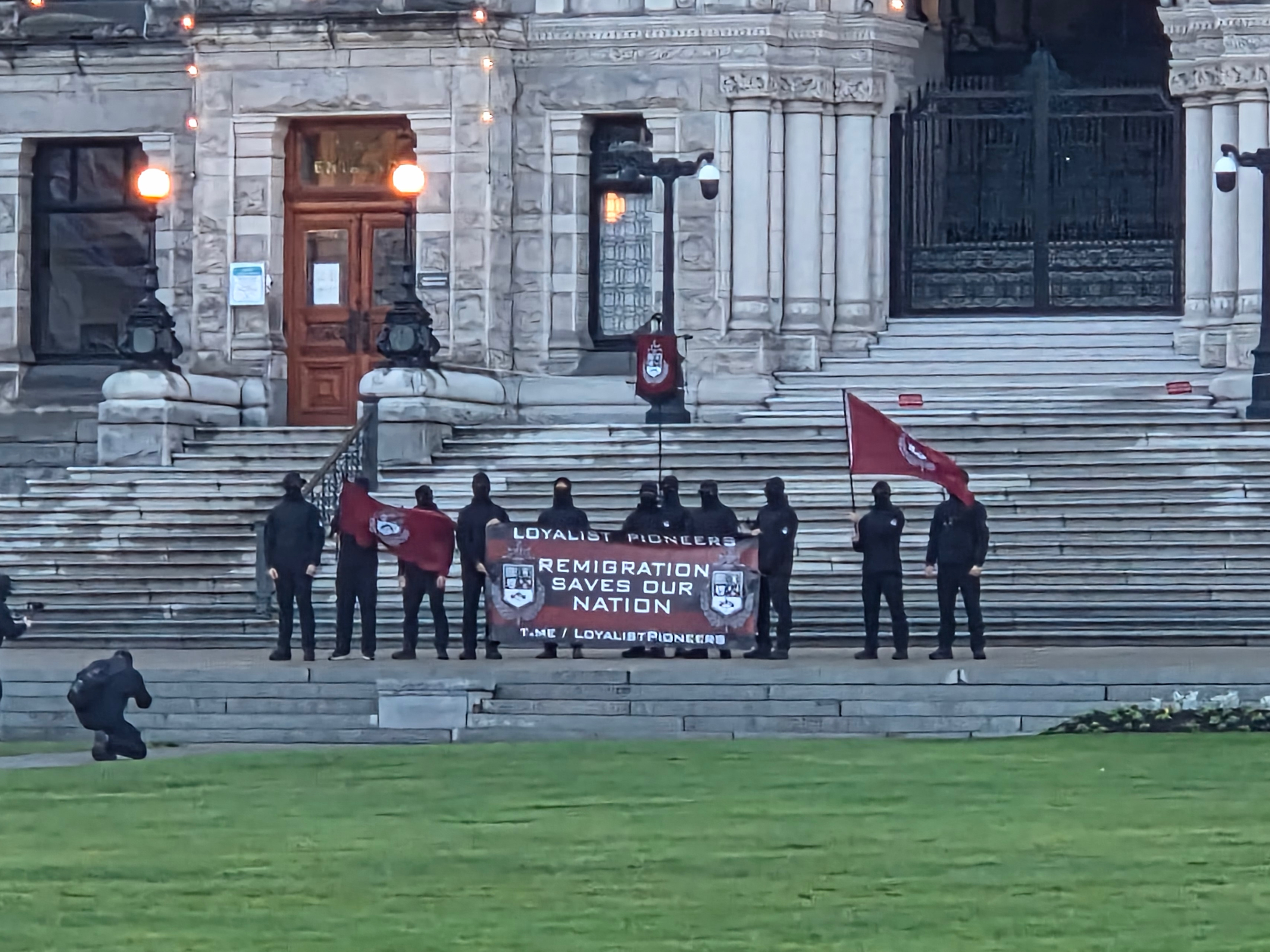 A group of men wearing black clothing and black masks stand in front of the BC legislature building, holding a maroon banner that says &ldquo;remigration now&rdquo; and &ldquo;loyalist pioneers.&rdquo;