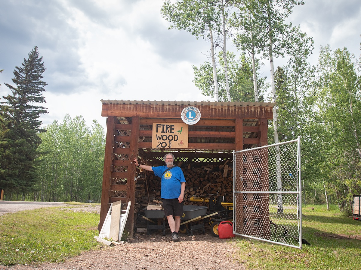 A man with greying hair and beard, wearing shorts and a T-shirt, stands in a small shed where a sign above the door reads, &lsquo;Firewood $20.&rsquo;