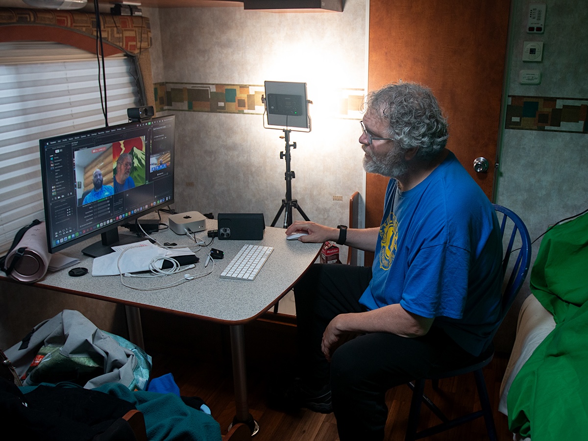 A man with greying hair and beard, wearing glasses and a blue T-shirt, sits at a computer and speaks with someone on the monitor.