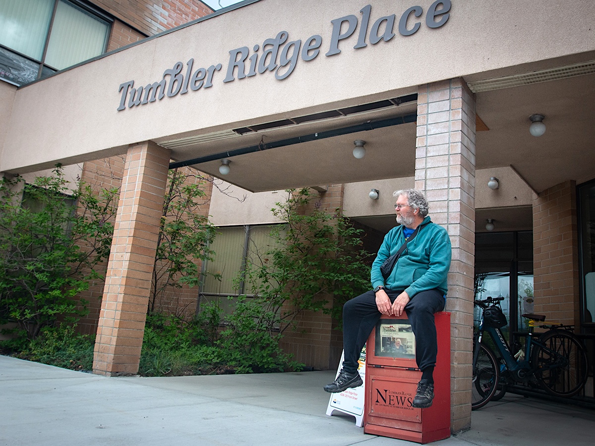 A man with grey hair and beard sits on an old, red newspaper box and looks into the distance. A sign over his head reads, &lsquo;Tumbler Ridge Place.&rsquo;