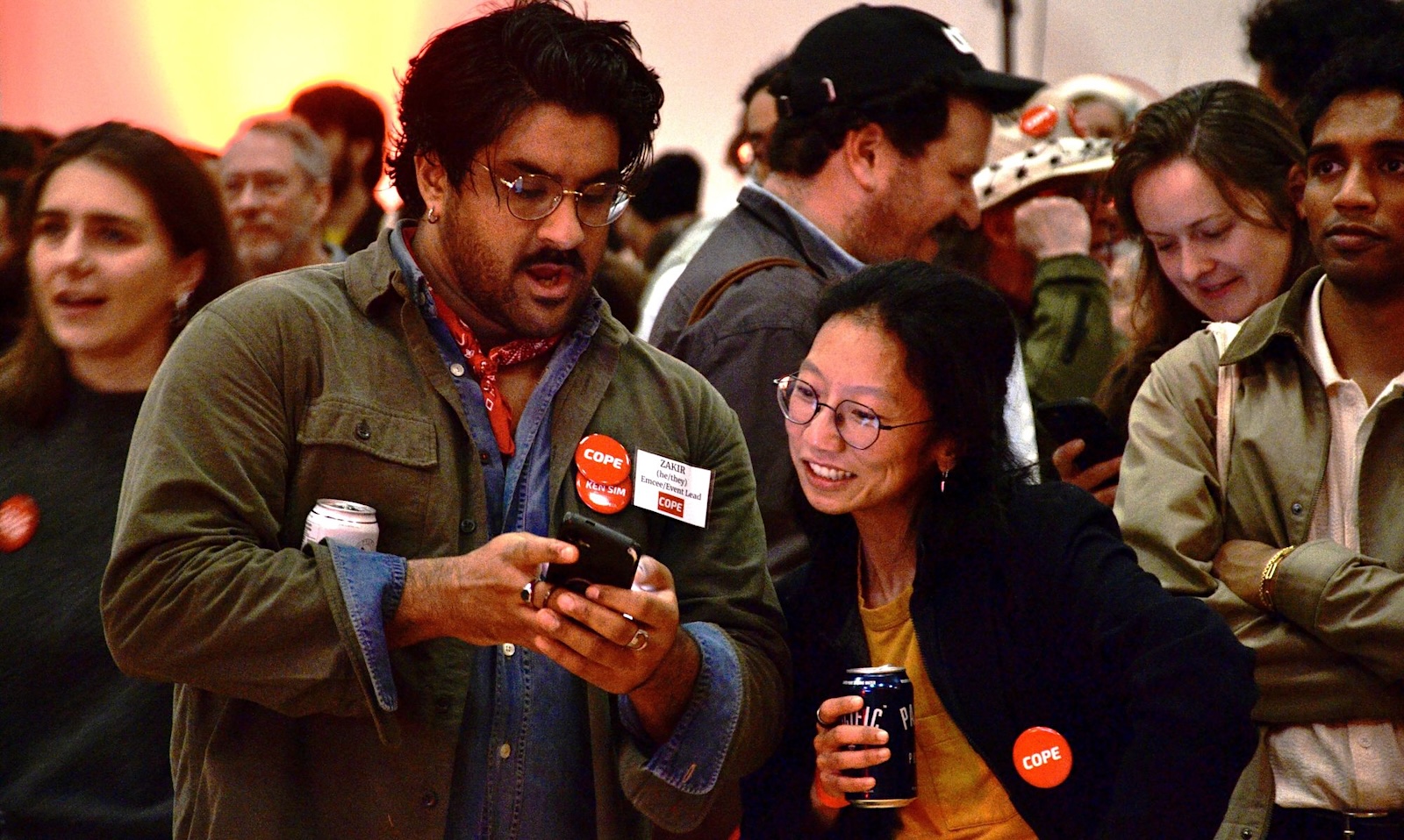 Amidst an animated crowd indoors, a South Asian young man with wide eyes looks at his phone, sharing the screen with a young Asian woman.