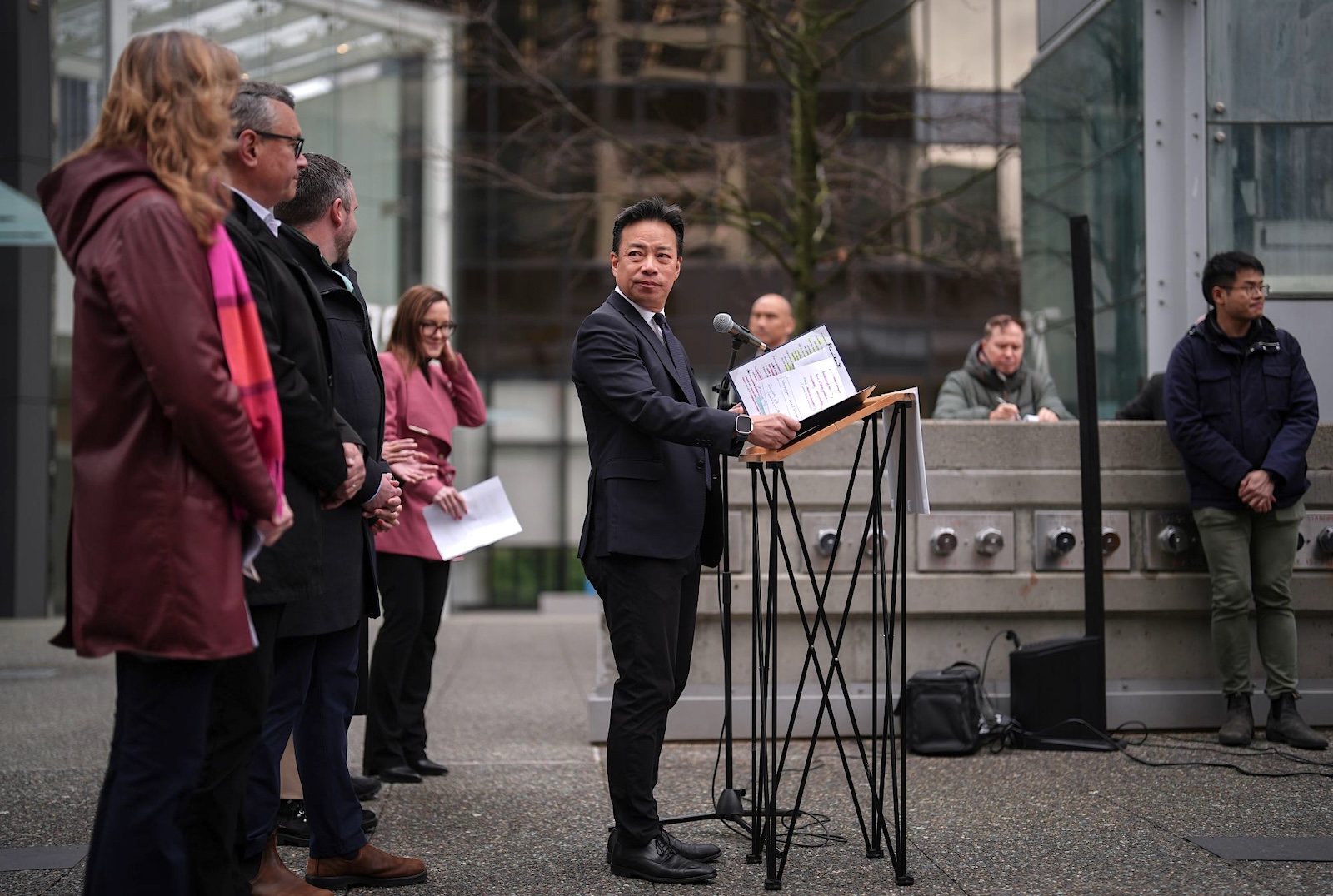 A young-middle-aged Chinese man stands at a lectern with microphones in an outdoors urban setting with people around him looking on.