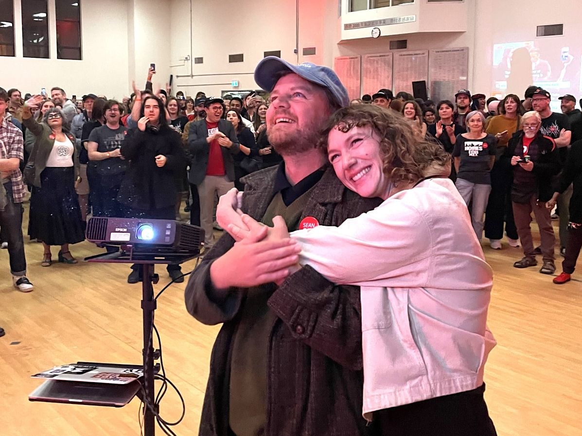 A man and a woman embrace while looking up at an unseen point. They&rsquo;re in a venue that looks like a gym with a crowd of people behind them.