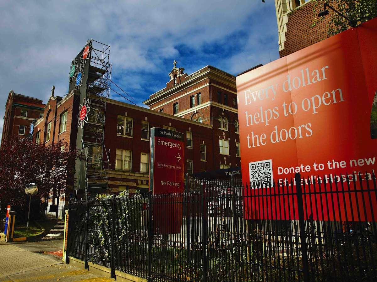A burgundy brick building is photographed against a bright blue sky. In the foreground to the right of the frame is a red sign that says, &lsquo;Every dollar helps to open the doors.&rsquo;