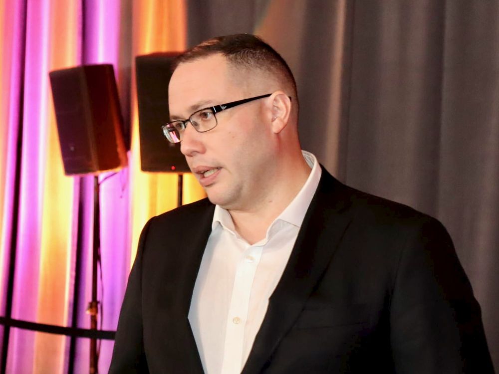A man with short dark hair and wide shoulders, wearing a white shirt and black blazer, speaks against a background of orange and purple curtains.
