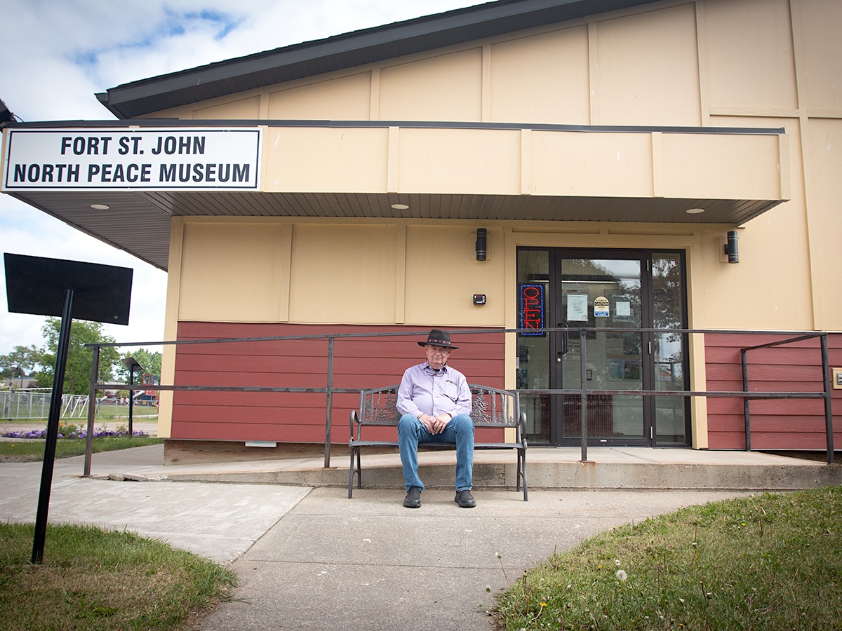 An older man wearing a black cowboy hat, purple shirt and jeans sits on a bench outside a building that bears the sign &lsquo;Fort St. John North Peace Museum.&rsquo;