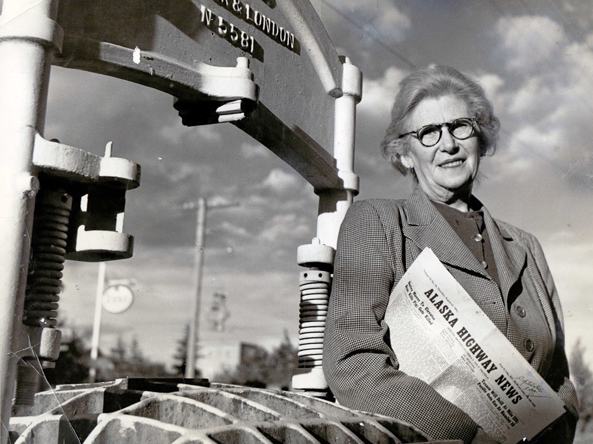 A black and white photo of a middle-aged woman with short grey hair and glasses holding a newspaper that says &lsquo;Alaska Highway News.&rsquo;