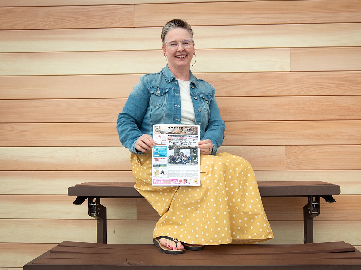 A woman with short hair and glasses wearing a denim jacket and yellow skirt sits on a bench and holds up a copy of a newsletter with the title &lsquo;Coffee Talk.&rsquo;