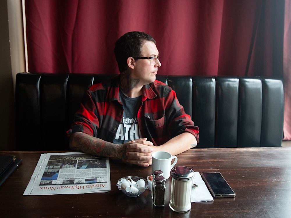 A man with short, dark hair and glasses sits at a table with his fingers clasped and looks out the window. On the table there is a cup of coffee and a newspaper with the title &lsquo;Alaska Highway News.&rsquo;