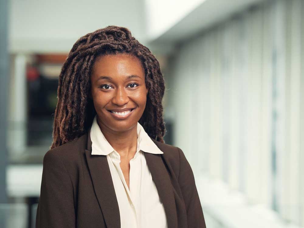 A 30-ish dark-skinned woman smiles at the camera. She has long dark hair and wears a brown jacket and white shirt.