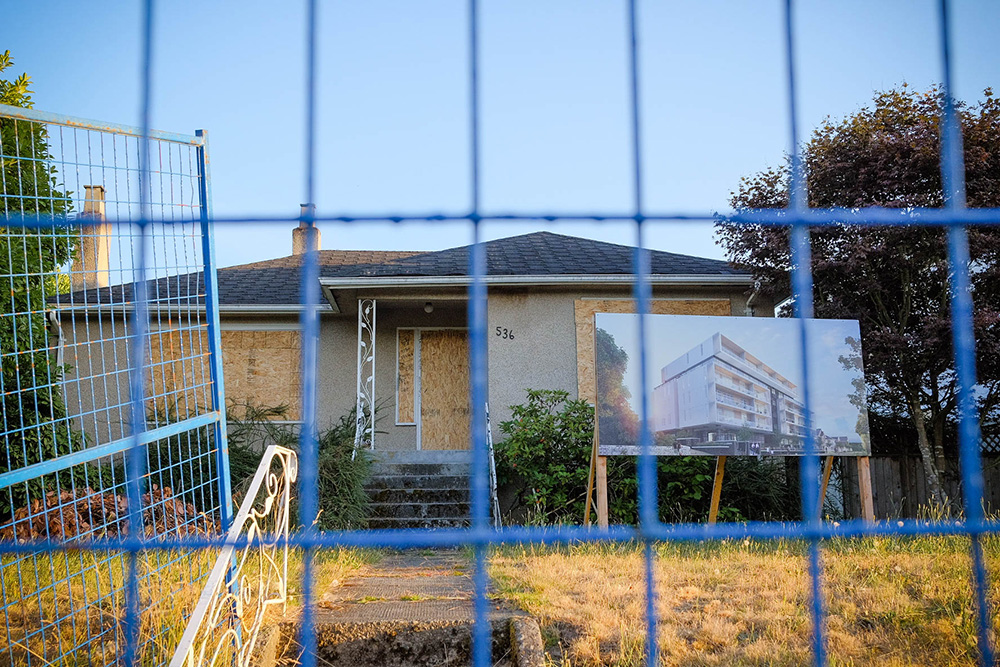 We’re looking through the grid of a blue fence towards a grey, derelict Vancouver bungalow with its windows boarded up. Beside it on a large wooden placard stands a portrait of the new high-rise development that will soon take its place.